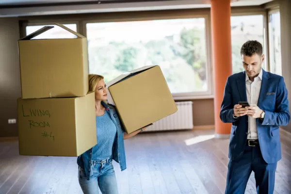 A woman carries two boxes labeled "Living Room #4" while a man checks his phone.