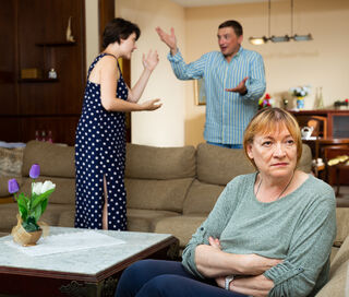 A woman sits with crossed arms while a couple argues in the background. A flower vase is on the table.