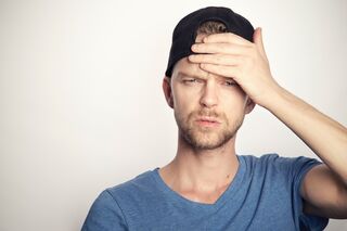 A man in a blue shirt and cap looks concerned, holding his forehead with a serious expression.