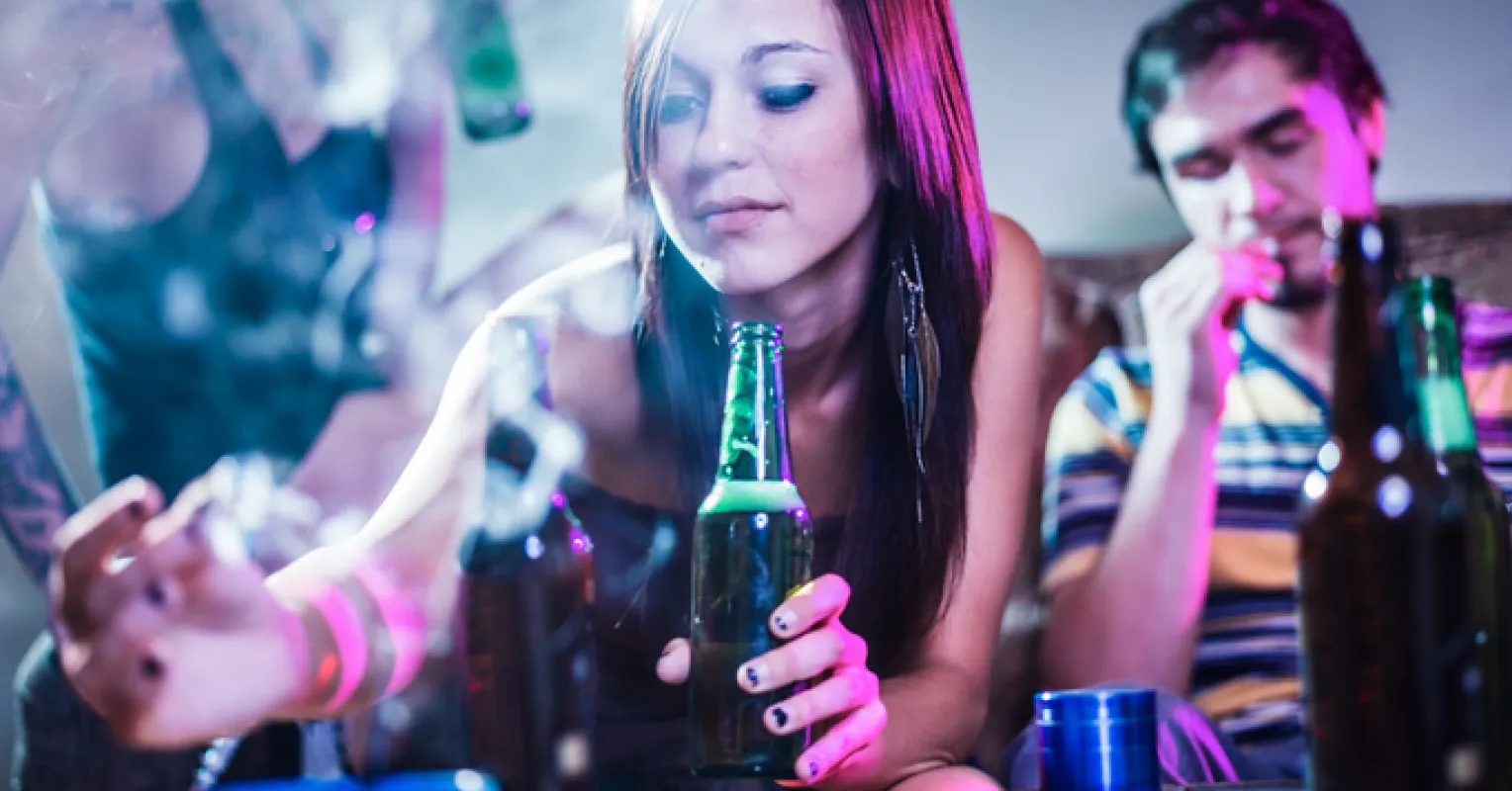 A young woman holds a green bottle, surrounded by others and smoke in a colorful, dimly lit setting.