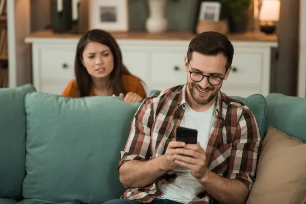 A man smiles at his phone while a woman looks at him with concern from behind.