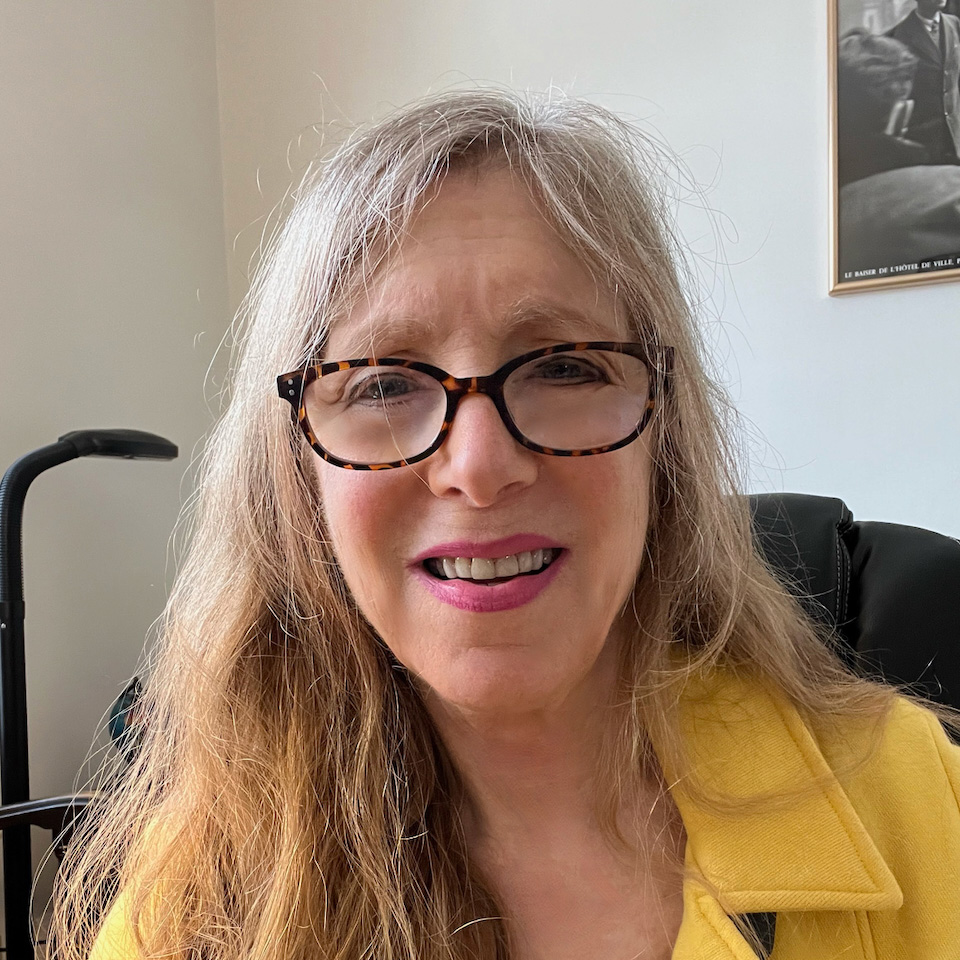 A woman with long hair and glasses smiles, wearing a yellow jacket in a well-lit room.