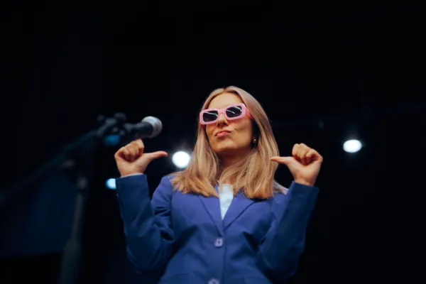 A woman in a blue suit and pink sunglasses poses confidently, pointing at herself on stage.
