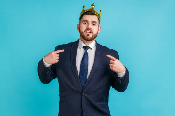 Man in a suit wears a crown and points to himself against a blue background.